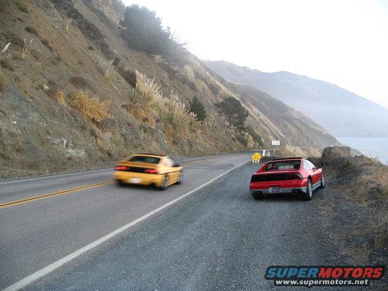 Red Pontiac Fiero parked on a coastal road, with a yellow Fiero driving away