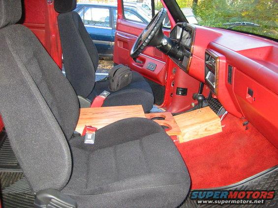 Interior of a red truck with black seats, wooden console, and dashboard. A blue car is visible through the open door