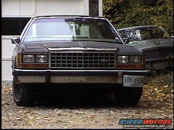 Front view of a dark Ford Crown Victoria with a license plate reading 100 1535. Another vehicle is partially visible behind it