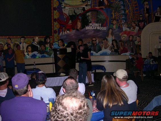 People gathered in front of a Planet Hollywood mural, with some standing on a stage and others seated in the foreground