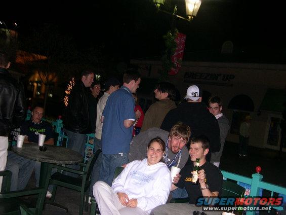 Group of people gathered outdoors at night, some seated at tables, others standing