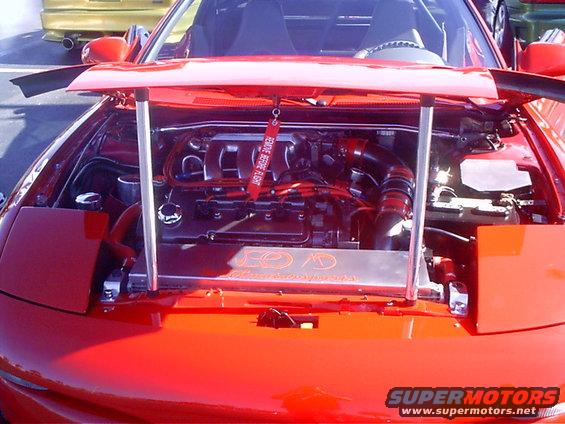 Red sports car engine bay with hood open, showing engine, intake, and radiator. A red 'REMOVE BEFORE FLIGHT' tag hangs from the engine