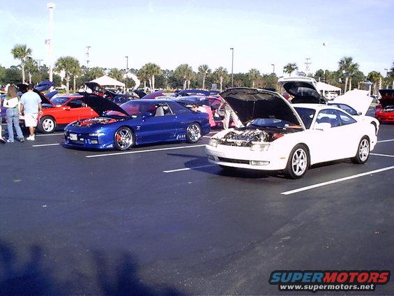 Several coupes with hoods open parked in a lot. One blue, one white, and one red car are visible