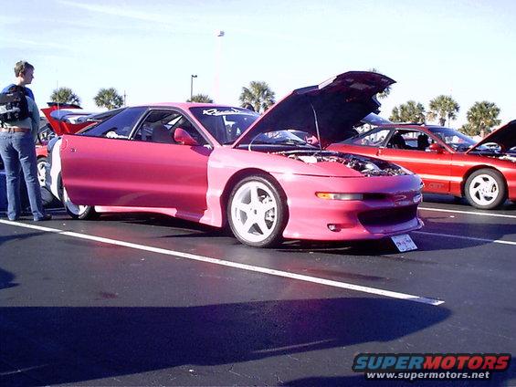 Pink sports coupe with hood and driver's door open, white wheels. Red sports coupe behind it