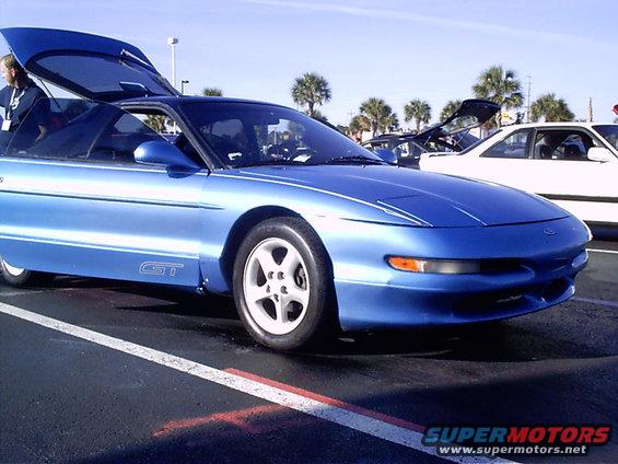 Blue 1995 Ford Probe GT coupe with doors and trunk open, parked on asphalt with palm trees in background