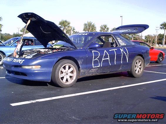 Blue coupe with hood open, guitar in engine bay, and 'BATN' painted on the side. Another car is visible in the background