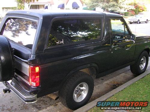 Black Ford Bronco parked on street, spare tire on back. Another white SUV is parked across the street
