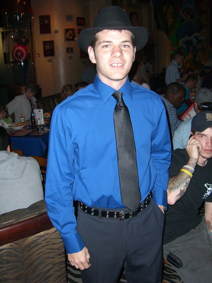 Man in blue shirt, black tie, and fedora stands in a crowded indoor setting