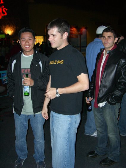 Three men stand in a dimly lit indoor space, one holding a beer bottle