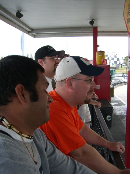 Several men watch a go-kart track, with a checkered flag visible in the background