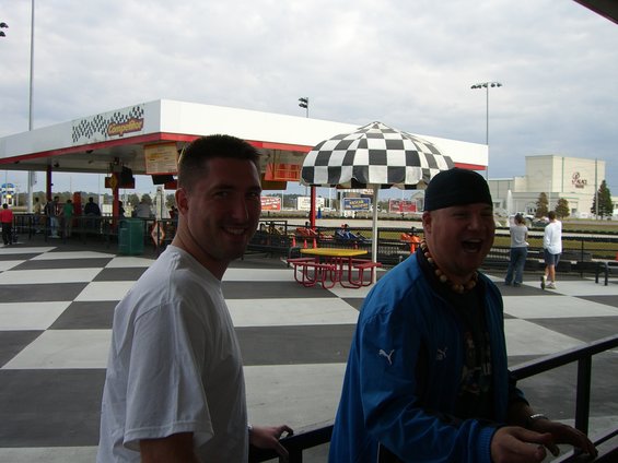 Two men smiling at a go-kart track with a checkered floor and a checkered umbrella