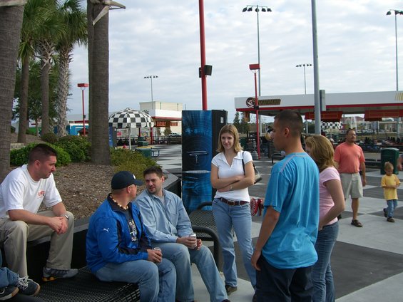 Group of people talking outdoors near palm trees and a gas station