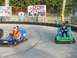 1996 Ford Probe - Two men race go-karts on an outdoor track, with a large billboard race car drivers in the background.