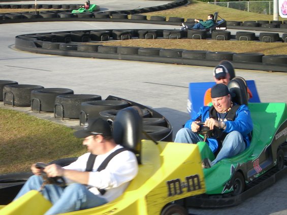 People racing go-karts on an outdoor track. One kart is yellow with M&M branding, another is green