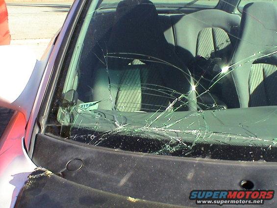 Cracked windshield of a car, showing spiderweb fractures and a small sticker that says TITAN