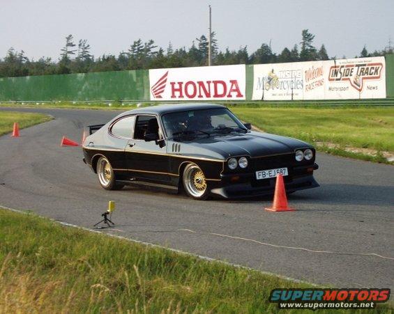Black coupe with gold trim on race track, orange cones visible