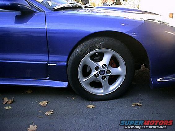 Blue 1993 Ford Probe front wheel and fender, showing a dent on the tire and some fallen leaves on the asphalt