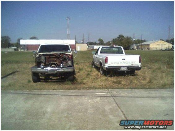 Front view of a damaged truck with exposed engine next to a white Chevrolet pickup