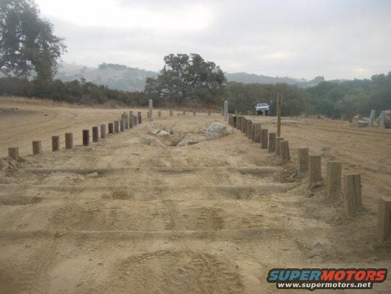 Dirt construction area with wooden posts and a white SUV in the distance