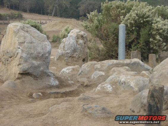 Rocky, sandy landscape with scattered boulders and dry brush under a hazy sky. A gray cylindrical marker stands among the rocks