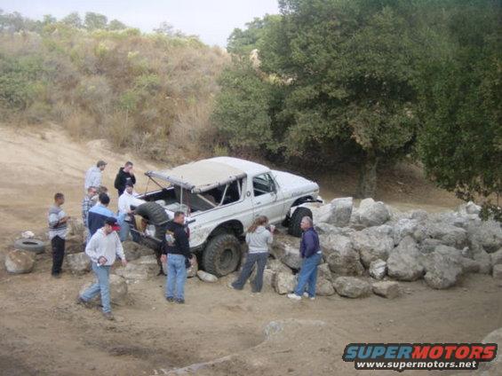 White off-road vehicle with a soft top, stuck on rocks, with people around it