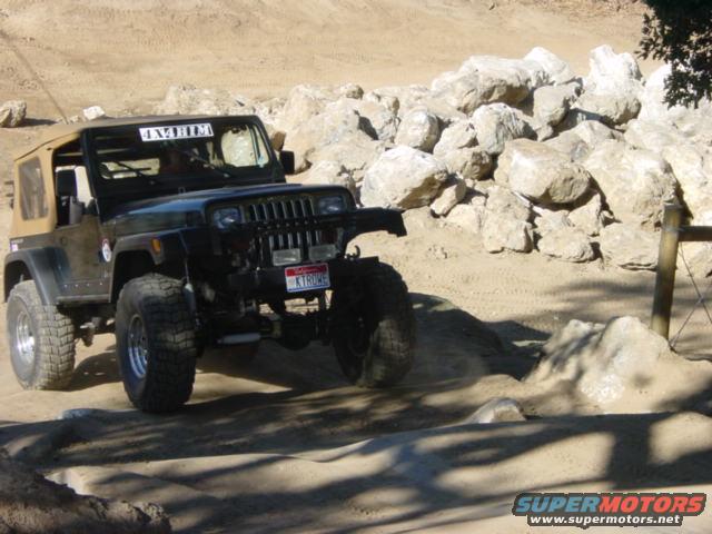 Black Jeep Wrangler TJ with tan soft top driving on a dirt trail with rocks