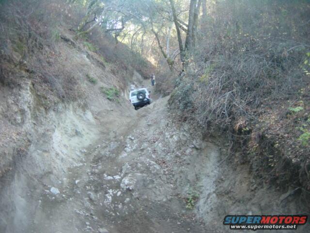 White SUV going down a steep, rocky trail with trees on the sides. Another person is visible further up the trail