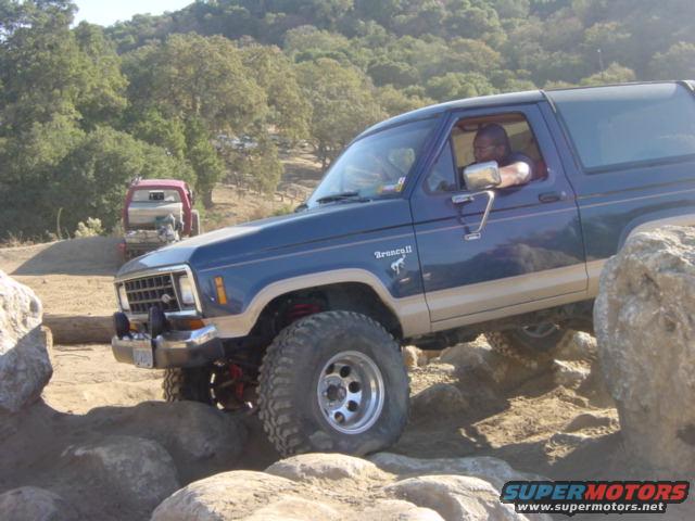 1986 Ford Bronco II navigating rocks on a trail, another red vehicle behind