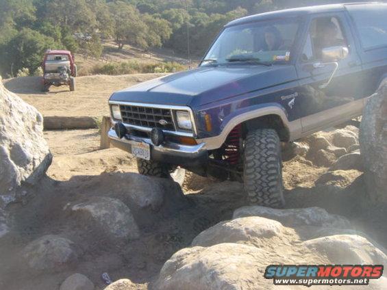 Blue 1986 Ford Bronco II navigating rocks on an off-road trail. Red truck in background