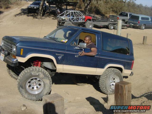 1986 Ford Bronco II with lifted suspension and large tires, driving off-road. Other vehicles visible in background