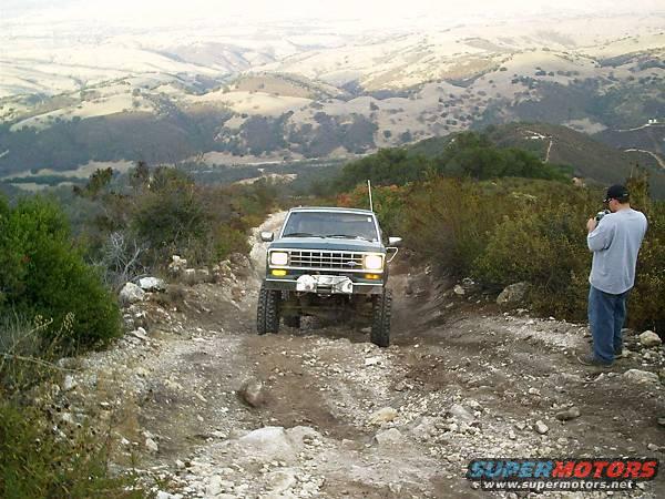 Dark green SUV with a winch on the front bumper driving up a rocky dirt road, with a man taking a photo on the right