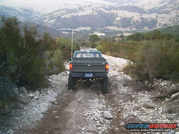 1986 Ford Bronco II driving on a rocky, off-road trail with hills in the background
