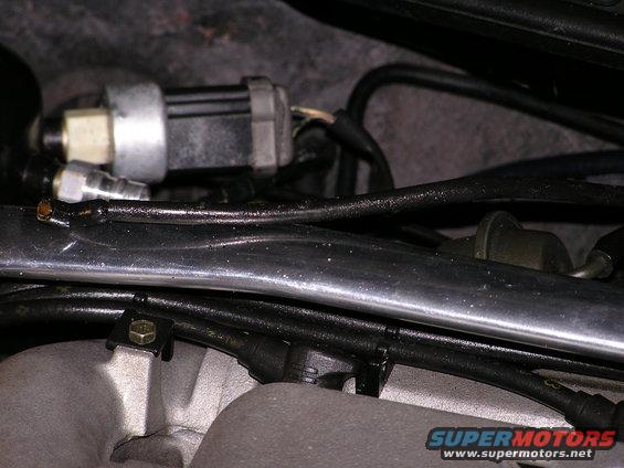 Close-up of a silver and black sensor with wires attached, mounted on a metal tube above engine parts