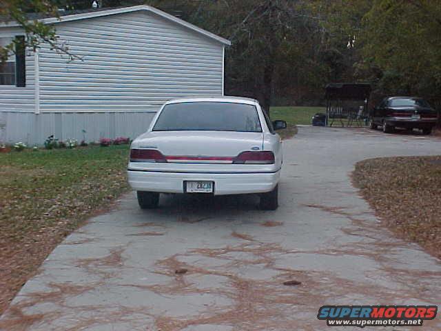 Rear view of a white sedan parked on a driveway, with a house on the left and another car in the background