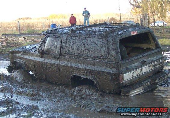 Mud-covered SUV stuck deep in mud with two people standing on its roof