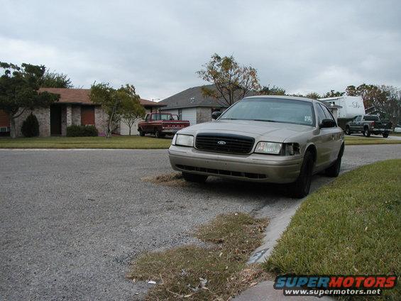 Tan Ford Crown Victoria parked on street, red pickup truck in driveway, another pickup truck near RV in background
