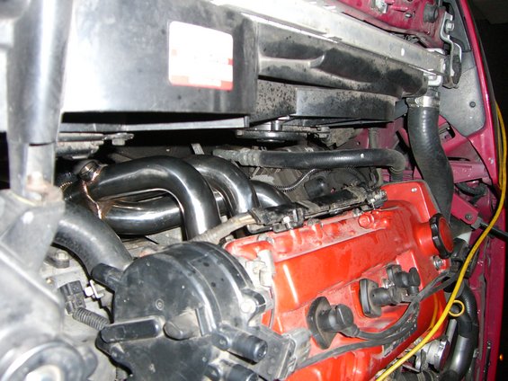 Engine bay with chrome exhaust headers and red valve cover. A black fan shroud is visible above