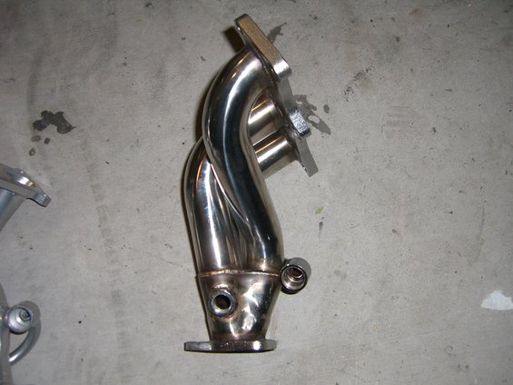 Shiny metal exhaust manifold with four pipes merging into one, resting on a concrete floor
