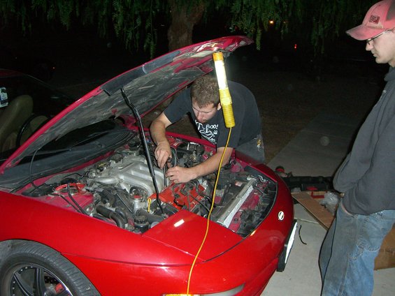 Red car with hood open, engine being worked on by a man. Another man stands nearby. Nighttime scene
