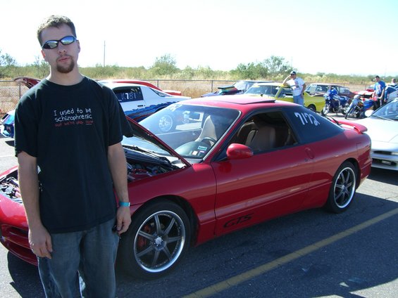guy in front of red coupe, hood open, other cars and motorcycles in background