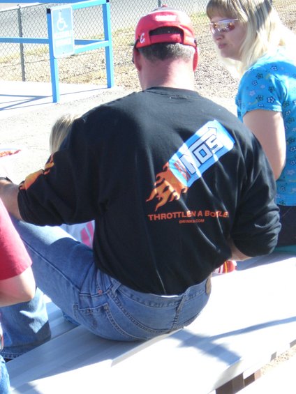 Man in black sweatshirt with "THROTTLE A BORE" graphic, sitting on bleachers with others