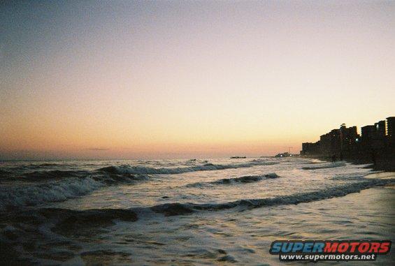 Ocean waves crash on the shore with a city skyline in the background during sunset. Watermark says SUPERMOTORS