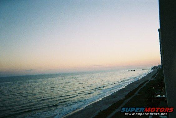 Ocean waves crash on a sandy beach during sunset, with a distant pier and buildings visible