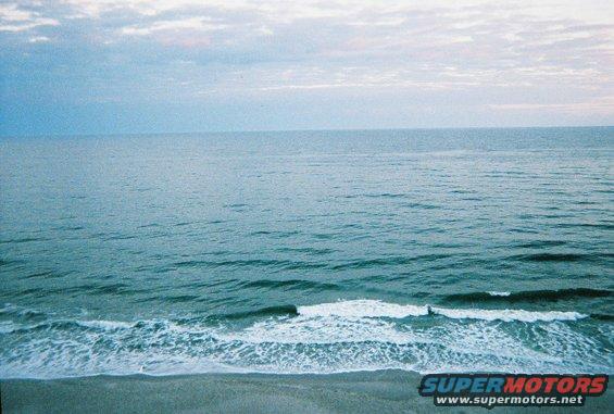 Ocean waves rolling onto a sandy beach under a cloudy sky. A small figure is visible in the water