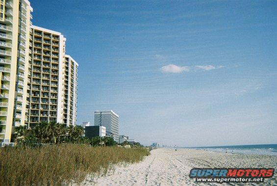 Beach scene with tall buildings on the left, ocean on the right, and a clear blue sky
