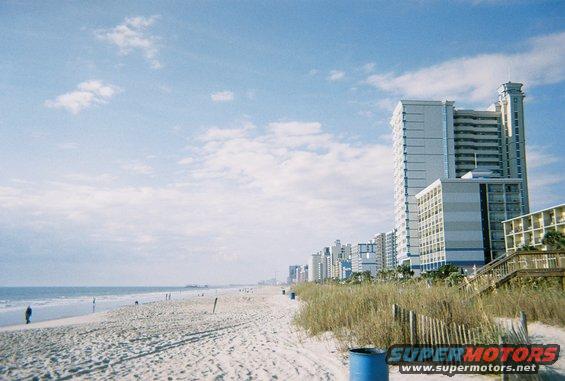Beach with ocean, sand, dunes, and hotels in the background. A few people are walking on the beach