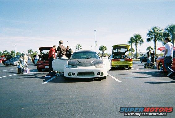 White sports car with carbon fiber hood and open doors, parked in a lot with other cars and people