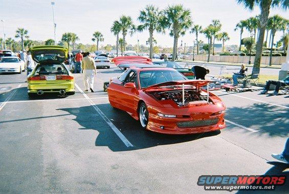 Red sports car with hood open, showing engine. Low front bumper, aftermarket wheels. Parked on asphalt
