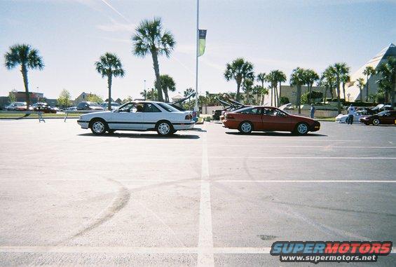 White coupe on left, red coupe on right, parked in a lot with palm trees and a pyramid-like building in the background