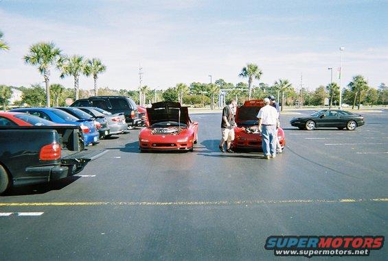 Several cars parked in a lot, some with hoods open. A dark coupe is parked to the right